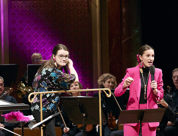 Aldo's Carnival family concert with conductor Zofia Kiniorska and saxophonist and artist in residence with the Orchestre de la Suisse Romande Valentine Michaud, speaking in a microphone on the Victoria Hall stage.